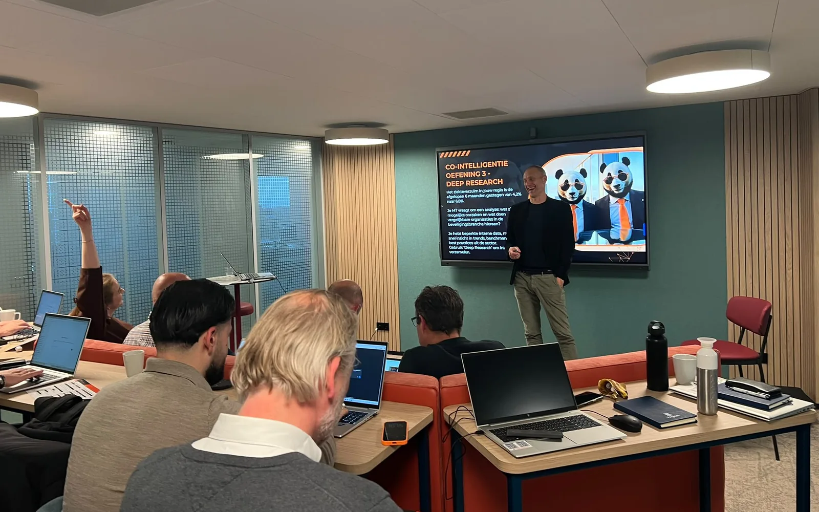 Een trainer van AI Panda staat voor een scherm met de oefening Deep Research; de deelnemers werken mee op hun laptop en een van hen steekt een hand op.